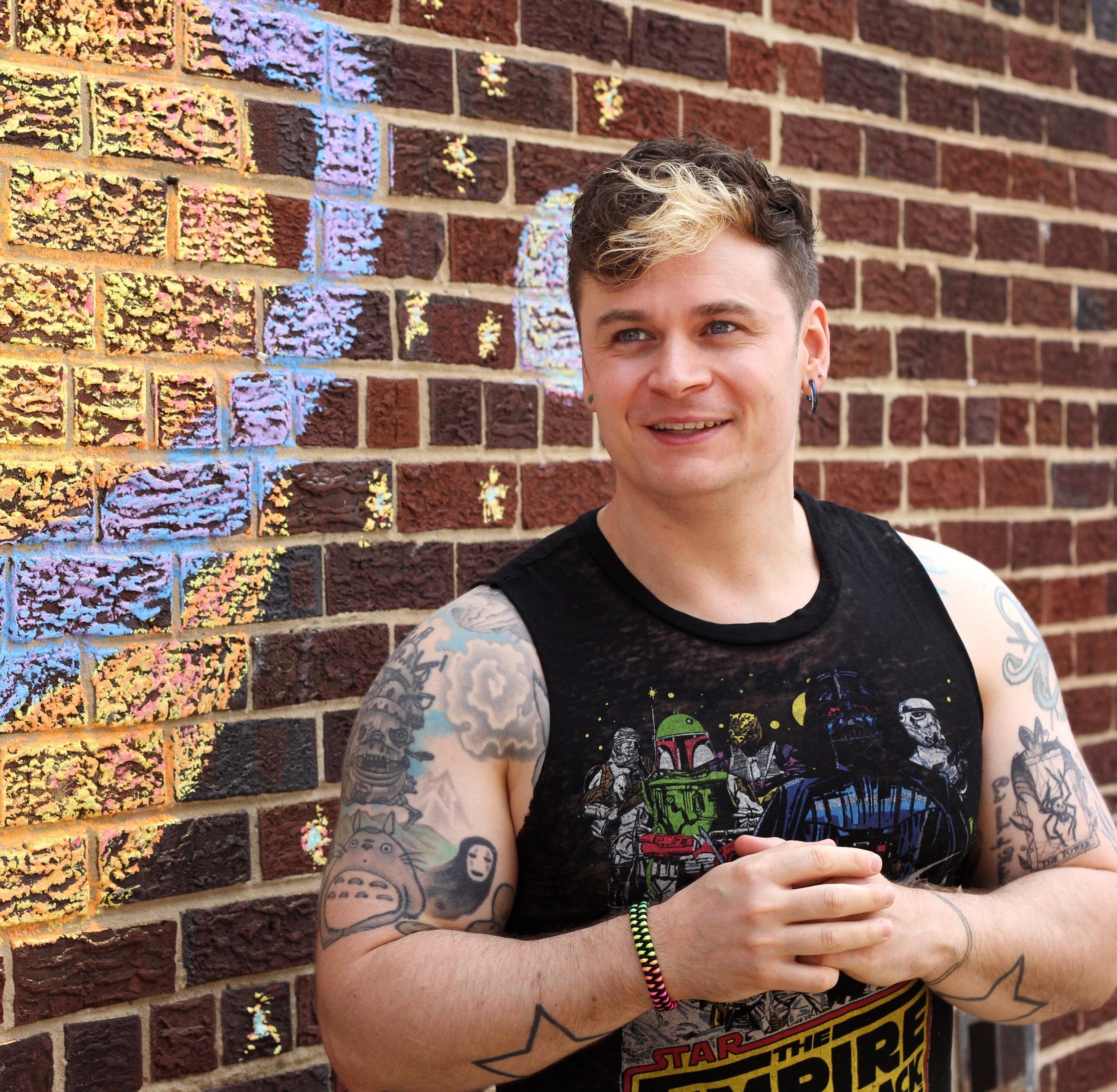 Trevor Stargazing In this photograph, Trevor, wearing a sleeveless Star Wars: The Empire Strikes Back shirt, stands against a red brick wall with a chalk drawing of stars and planets on it as he stares off into the distance and smiles.