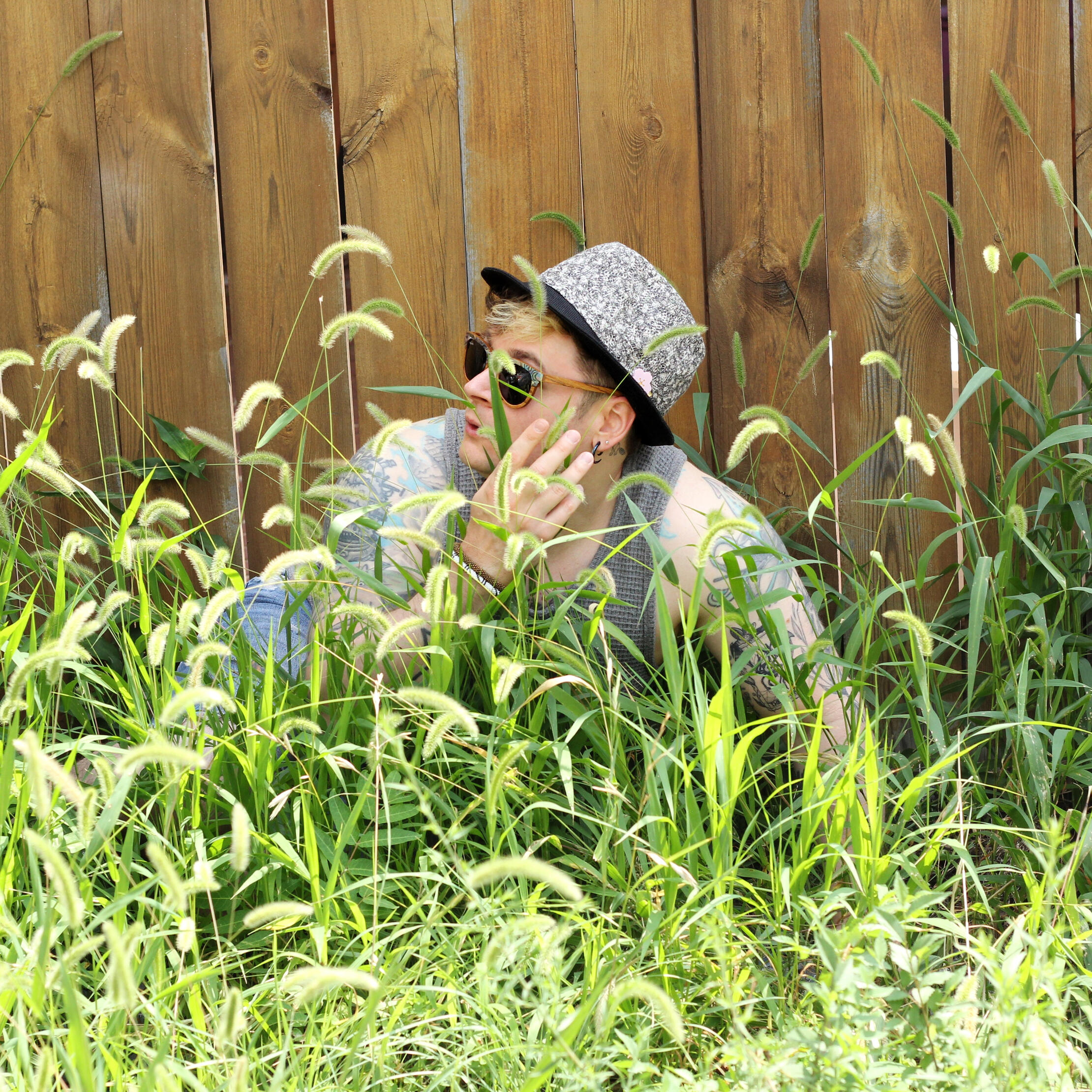 Trevor Hides from Predators Trevor Garner, wearing some fancy wood framed sunglasses, hand made blue-grey fedora, and a waffled grey tank top, hides behind tall grass against a an old wooden fence.