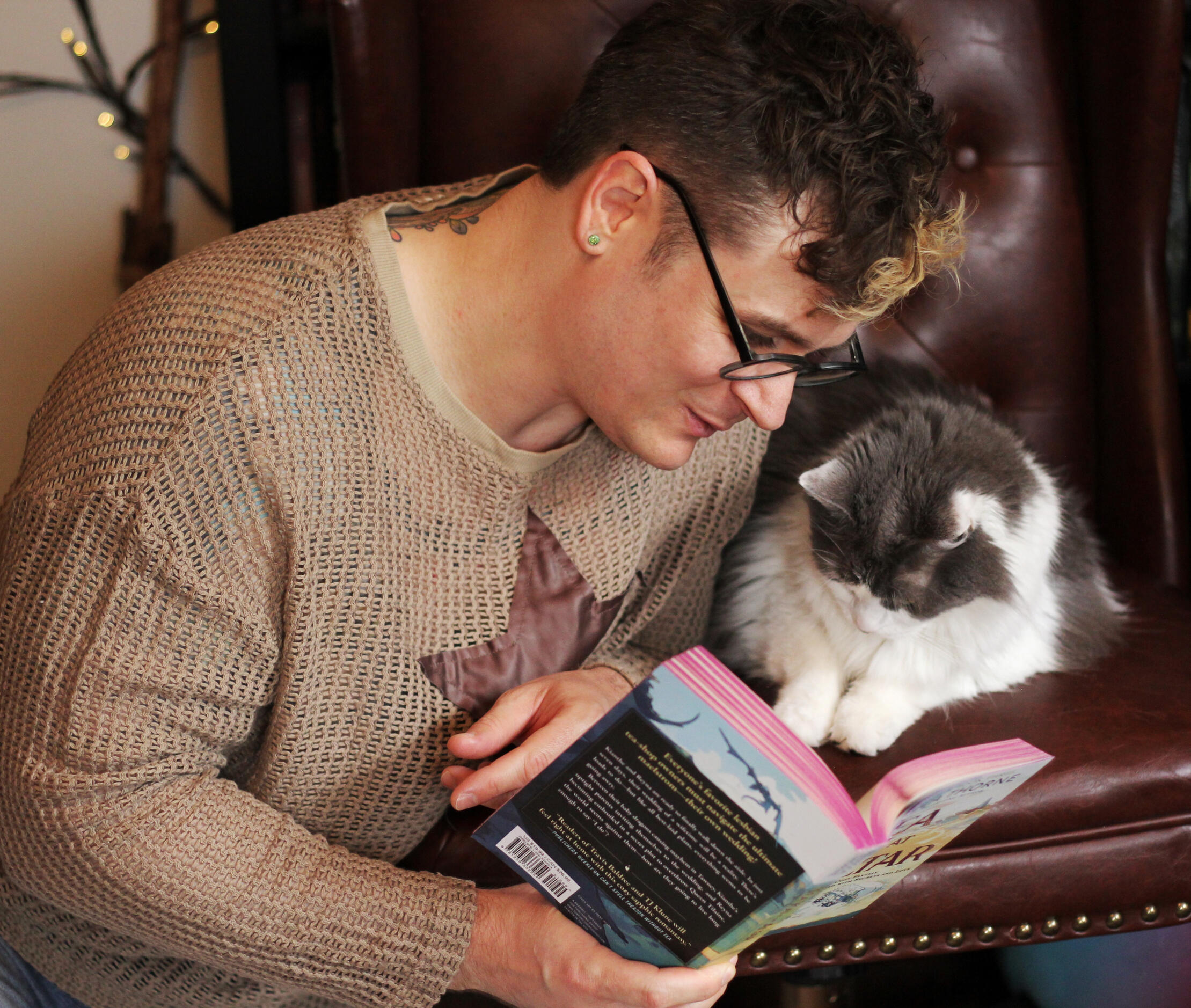 Trevor and Sophie Trevor Garner, in glasses and a fishnet tan long sleeved shirt with a leather star sewn onto the front, kneels as he reads one of his favorite books to his grey and white long haired cat Sophie, who sits beside him in a chair and seems very interested in t